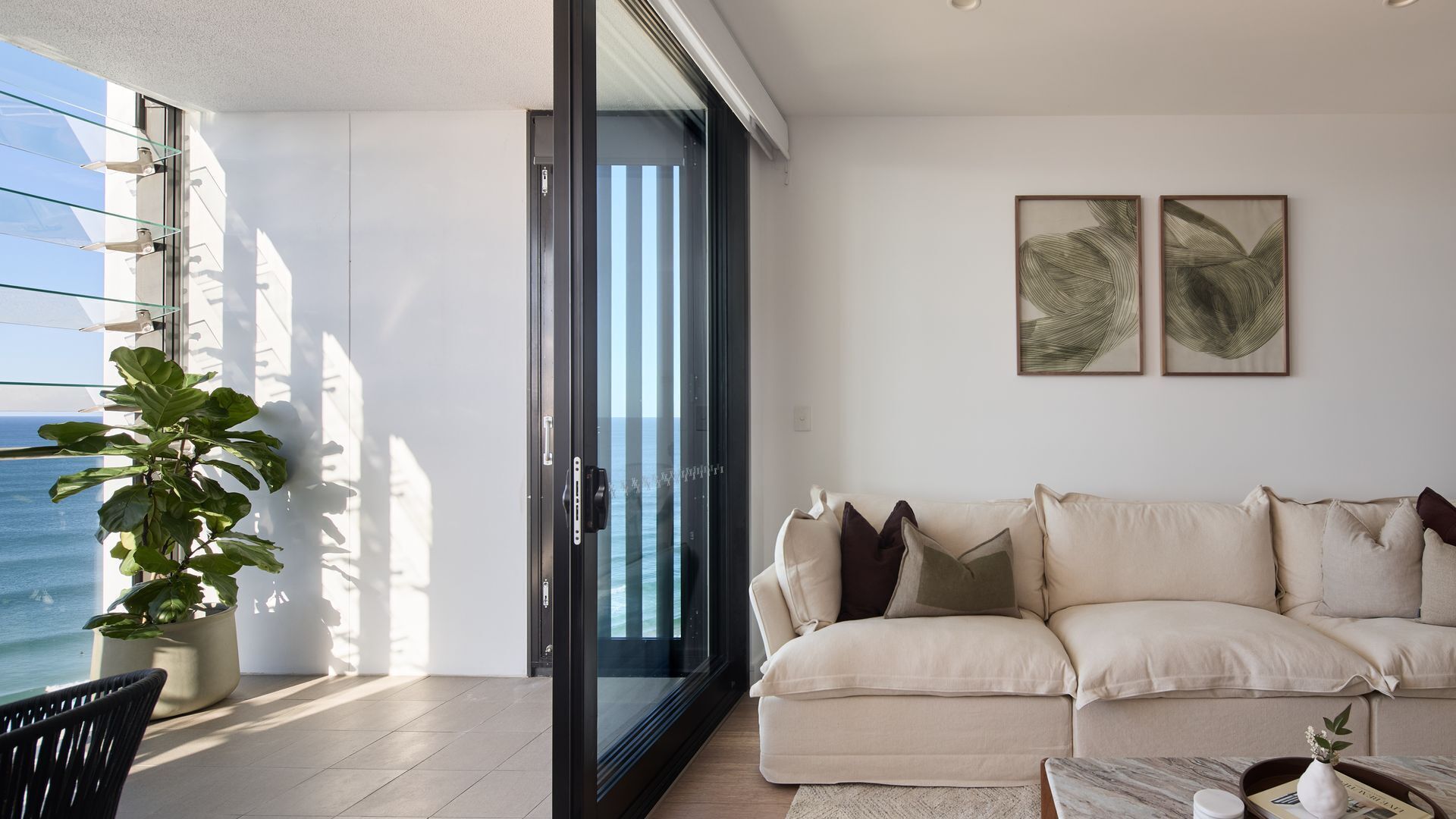 A modern living room with a sofa, a potted plant, and a view of the sea through a glass door.