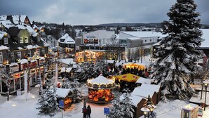 Une place de ville enneigée, avec un carrousel, des boutiques et un grand sapin de Noël.