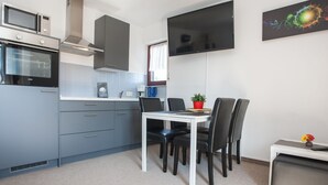 Une cuisine compacte équipée d’un four à micro-ondes, d’une cuisinière et de meubles de rangement modernes. Une table à manger avec des chaises est placée près d’un grand téléviseur à écran plat fixé au mur.