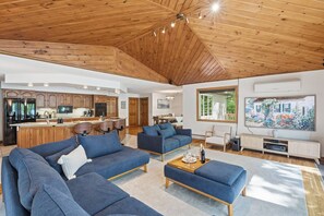 Een ruime woonkamer met een houten plafond, een keuken met houten kastjes en een groot raam met uitzicht op groen.