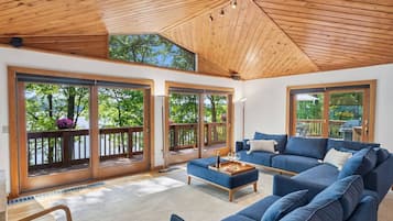 Un vaste espace de vie agrémenté d’un canapé bleu, d’un plafond en bois et de grandes portes-fenêtres coulissantes donnant sur un balcon avec vue sur les arbres et l’eau.