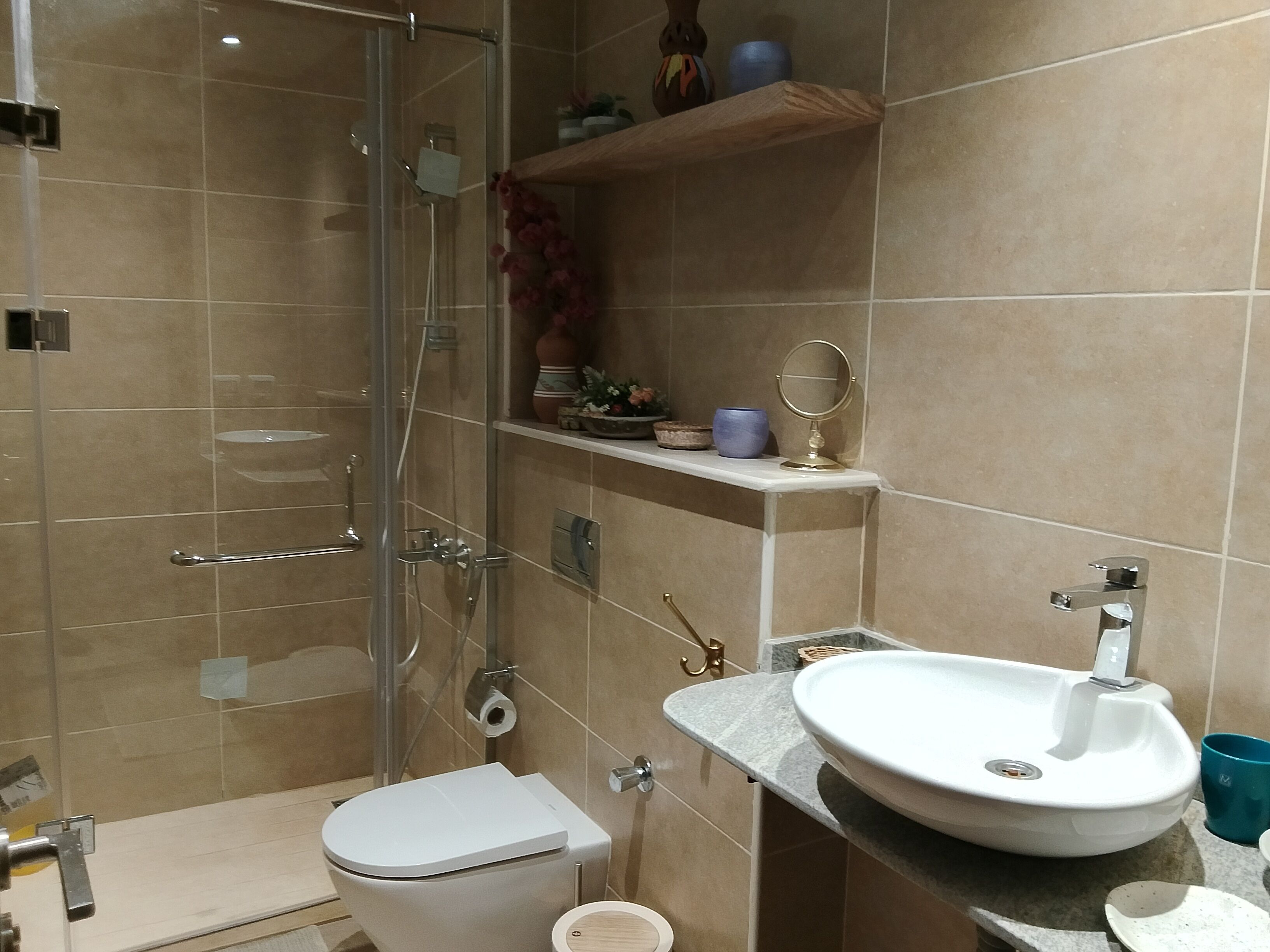 A modern bathroom with a glass shower enclosure, a white toilet, a sink with a marble countertop, and a shelf with decorative items.