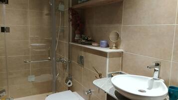 A modern bathroom with a glass shower enclosure, a white toilet, a sink with a marble countertop, and a shelf with decorative items.