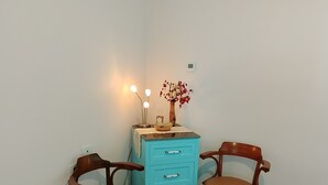 Una habitación con dos sillas de madera, un aparador azul con cajones, una mesita con un florero y una lámpara fijada a la pared.