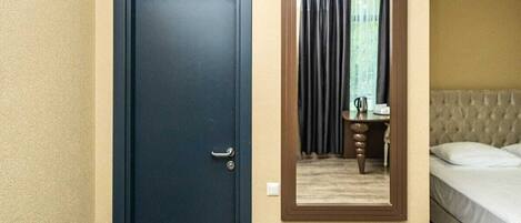 Habitación de hotel con una puerta azul oscuro, un pequeño banco de madera, una cama con ropa blanca y un espejo en la pared.