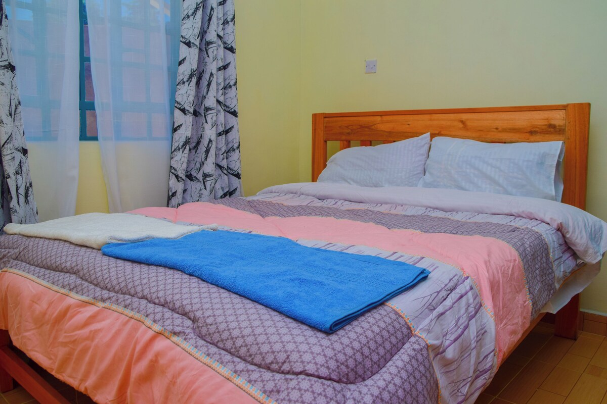 Una cama con varias mantas y almohadas, cabecera de madera y una ventana con cortinas traslúcidas.