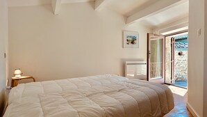 Ein Schlafzimmer mit einem großen Bett, einem Nachttisch mit Lampe, einem gerahmten Bild an der Wand und einer Tür zu einem Außenbereich.