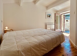 Ein Schlafzimmer mit einem großen Bett, einem Nachttisch mit Lampe, einem gerahmten Bild an der Wand und einer Tür zu einem Außenbereich.