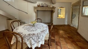 Ein Essbereich mit einem runden Tisch, bedeckt mit einer gemusterten Tischdecke, umgeben von Holzstühlen. Im Hintergrund ist ein Kamin mit Sims erkennbar, und ein Spiegel hängt an der Wand.
