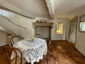 Ein Essbereich mit einem runden Tisch, bedeckt mit einer gemusterten Tischdecke, umgeben von Holzstühlen. Im Hintergrund ist ein Kamin mit Sims erkennbar, und ein Spiegel hängt an der Wand.