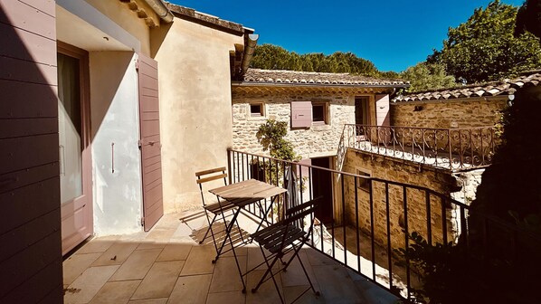 Un balcon avec une table et des chaises, un bùtiment en pierre avec un toit en tuiles et un ciel bleu dégagé.