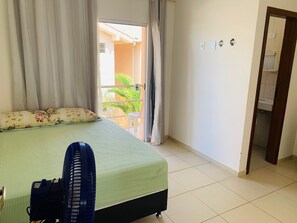 Quarto com cama, ventilador, janela com cortinas e vista para uma varanda com plantas.