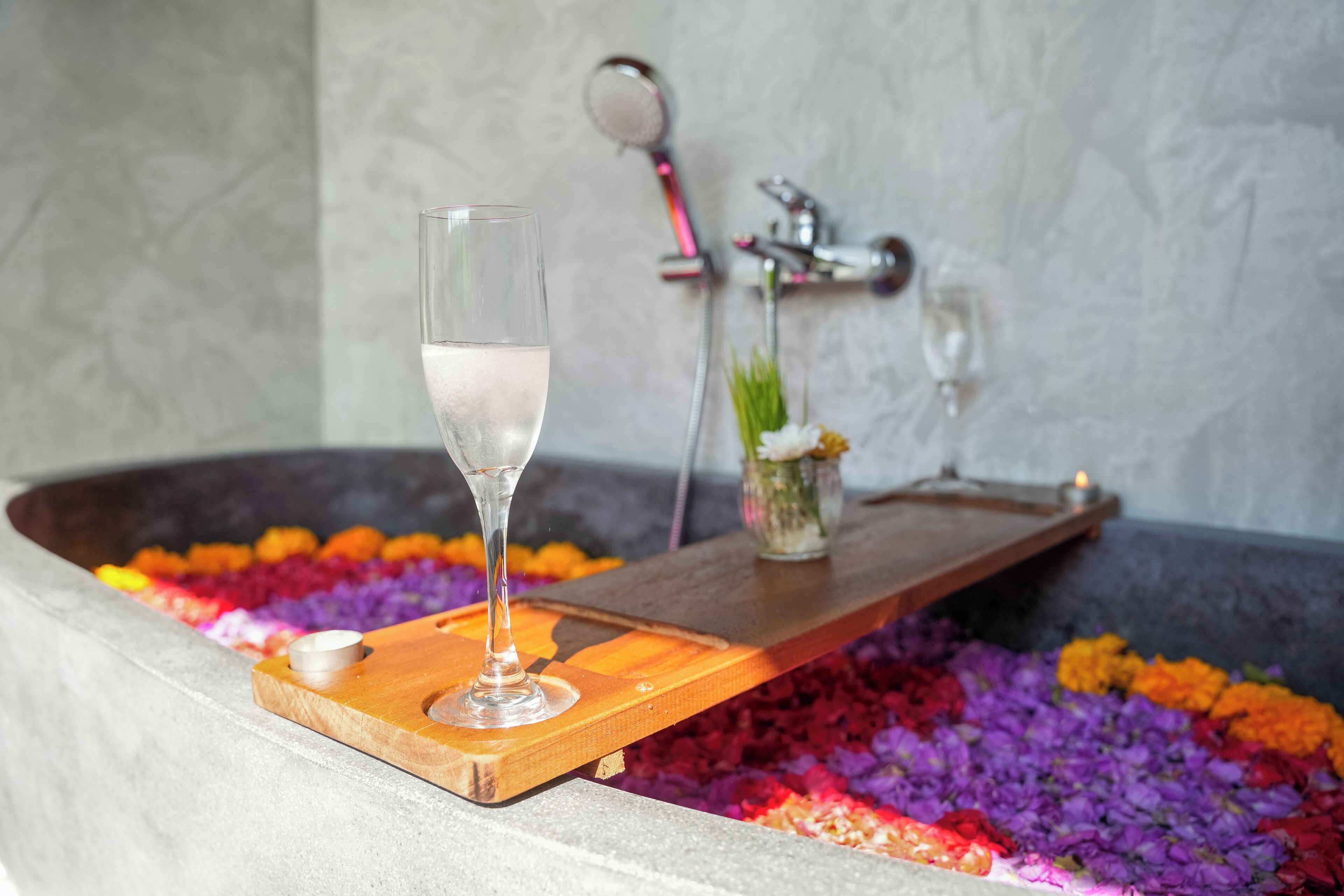 A bathtub filled with colorful flower petals, a glass of water, and a wooden tray.