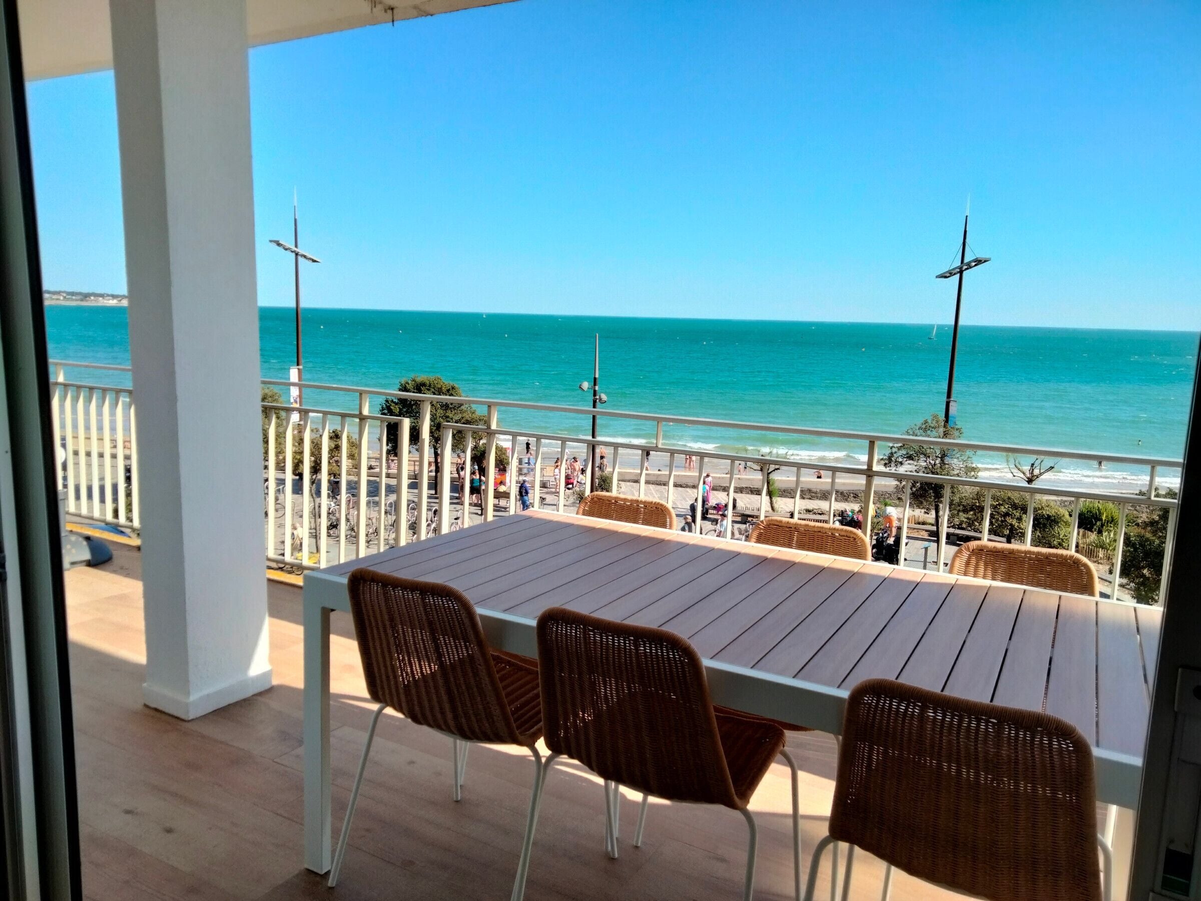 Un balcon avec une table en bois et des chaises en osier, donnant sur une plage animée et un quai.