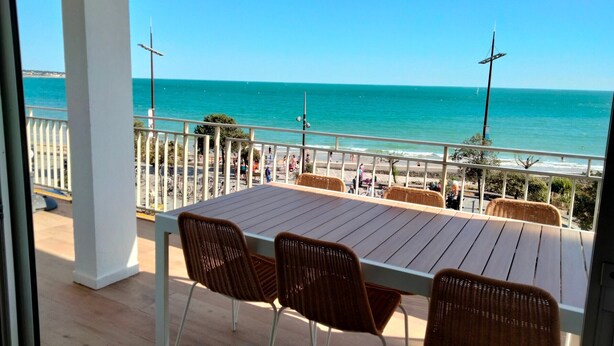 Ein Balkon mit Holztisch und Korbstühlen mit Blick auf einen Strand, an dem sich Menschen befinden und eine Mole liegt.