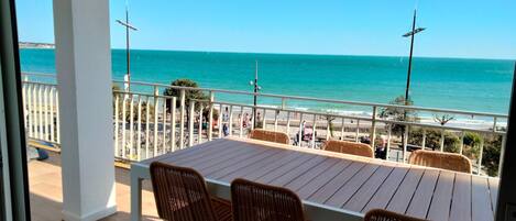 Un balcon avec une table en bois et des chaises en osier, donnant sur une plage animée et un quai.