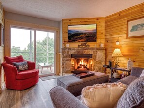 Ein gemütliches Blockhauss-Interieur mit Kamin aus Stein, einem roten Sessel und einem großen Fenster mit Blick auf Grünflächen.