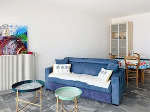 A living room with a blue sofa, a glass coffee table, a side table, a radiator, and a colorful abstract painting. - Beautiful apartment with sea view - 4 guests (Les Sables-d'Olonne)