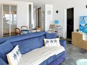 A living room with a blue sofa, a dining table with chairs, a white cabinet, and a painting on the wall. - Beautiful apartment with sea view - 4 guests (Les Sables-d'Olonne)