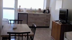 Een kleine keuken met een eettafel, stoelen, een kleine koelkast, een televisie en een raam met jaloezieën.
