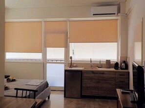 A compact hotel room with a bed, a small desk, a kitchenette, and a window with blinds.