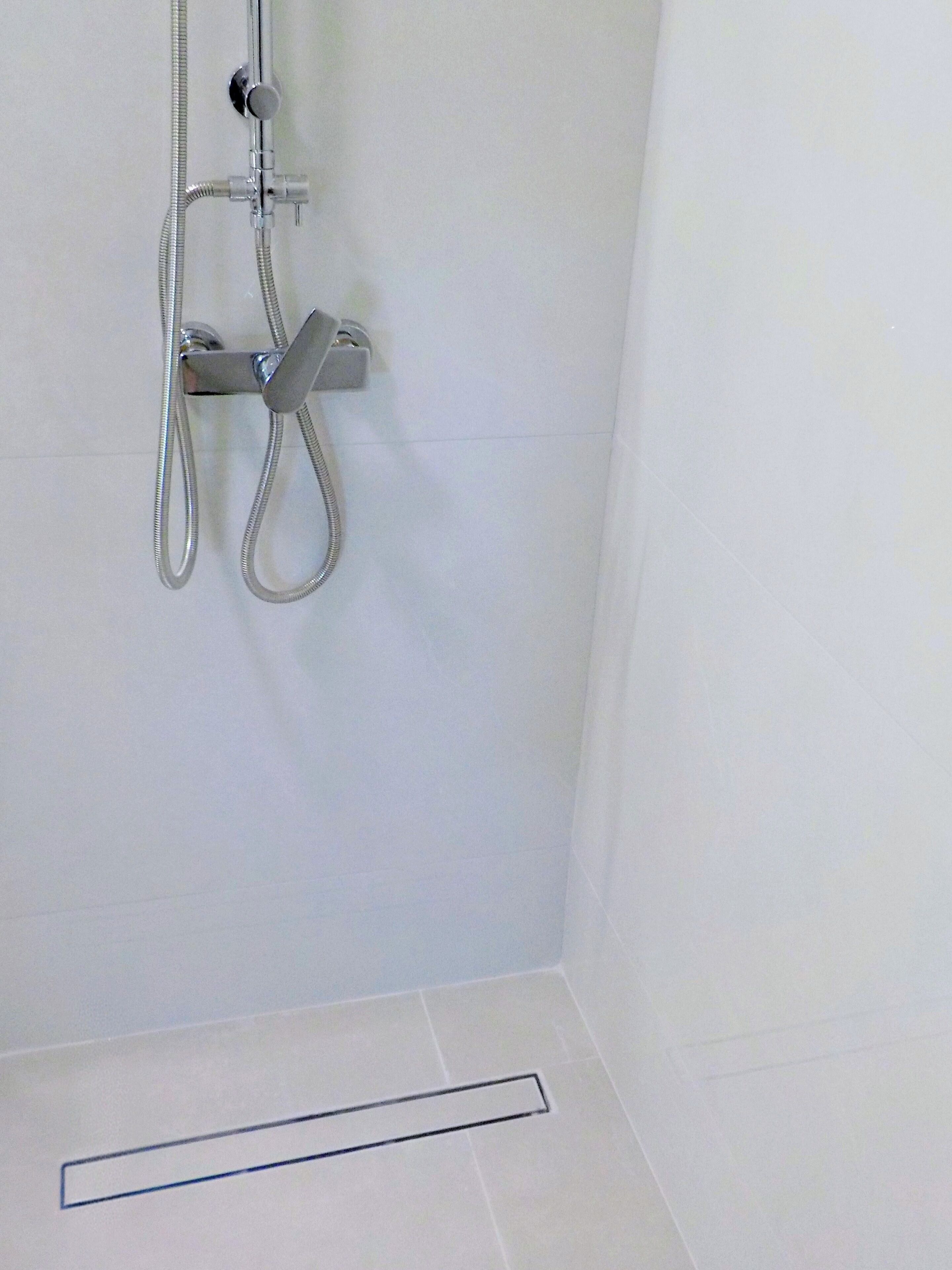 Une douche équipée d’un flexible et d’un pommeau de douche fixé au mur, ainsi qu’un sol carrelé et un drain.