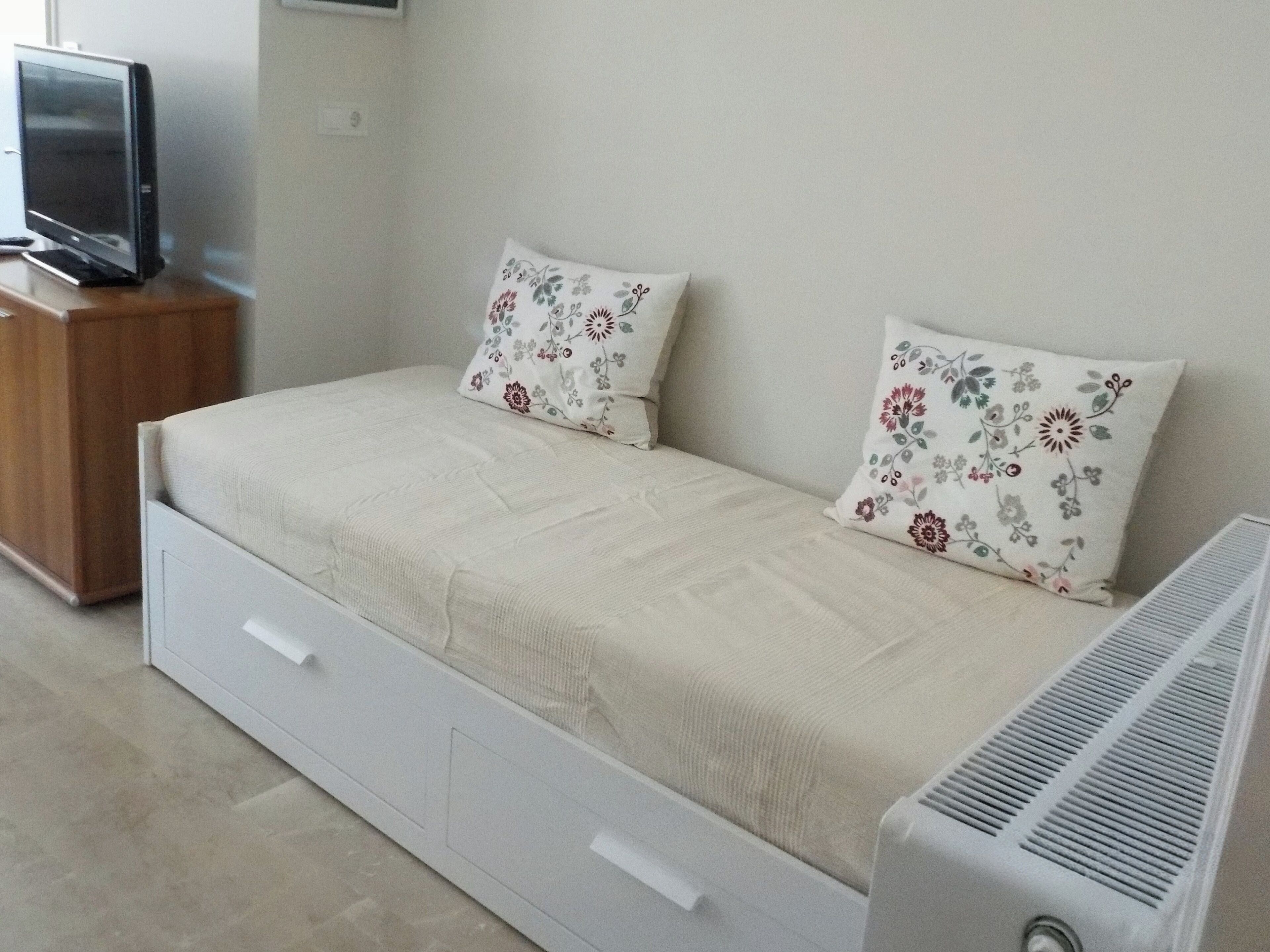 Een enkel bed met een witte matras en kussens met bloemenprint, een houten nachtkastje met een televisie, en een witte ladekast.