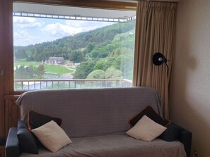 A room with a view of a green landscape through a window, a sofa with cushions, and a lamp on the wall. - Comfortable studio, 4 guests, Super Besse center (Besse-et-Saint-Anastaise-Super Besse)