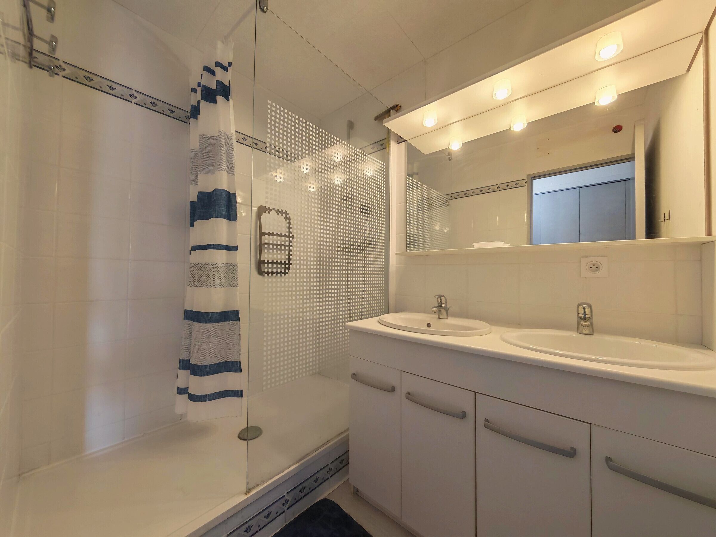 Un bany amb una cabina de dutxa de vidre, un lavabo blanc amb un mirall a sobre i una paret revestida de rajoles amb un disseny geomètric.