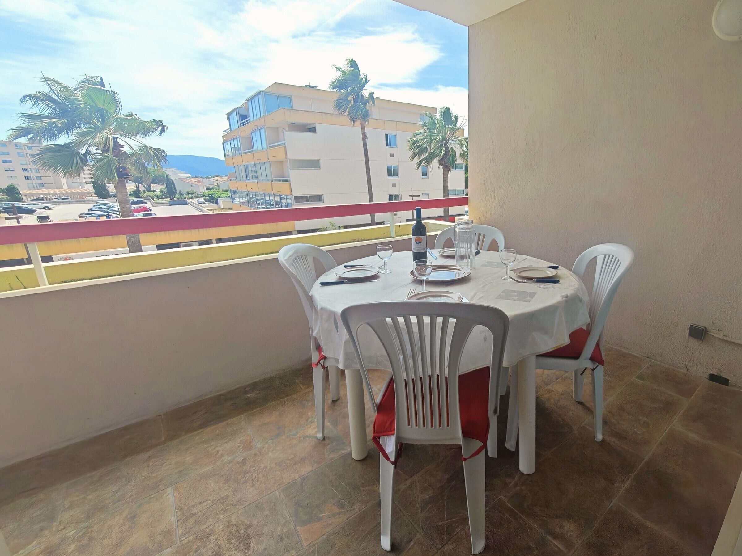 A balcony with a round table set for dining, white plastic chairs, and a view of buildings and palm trees.