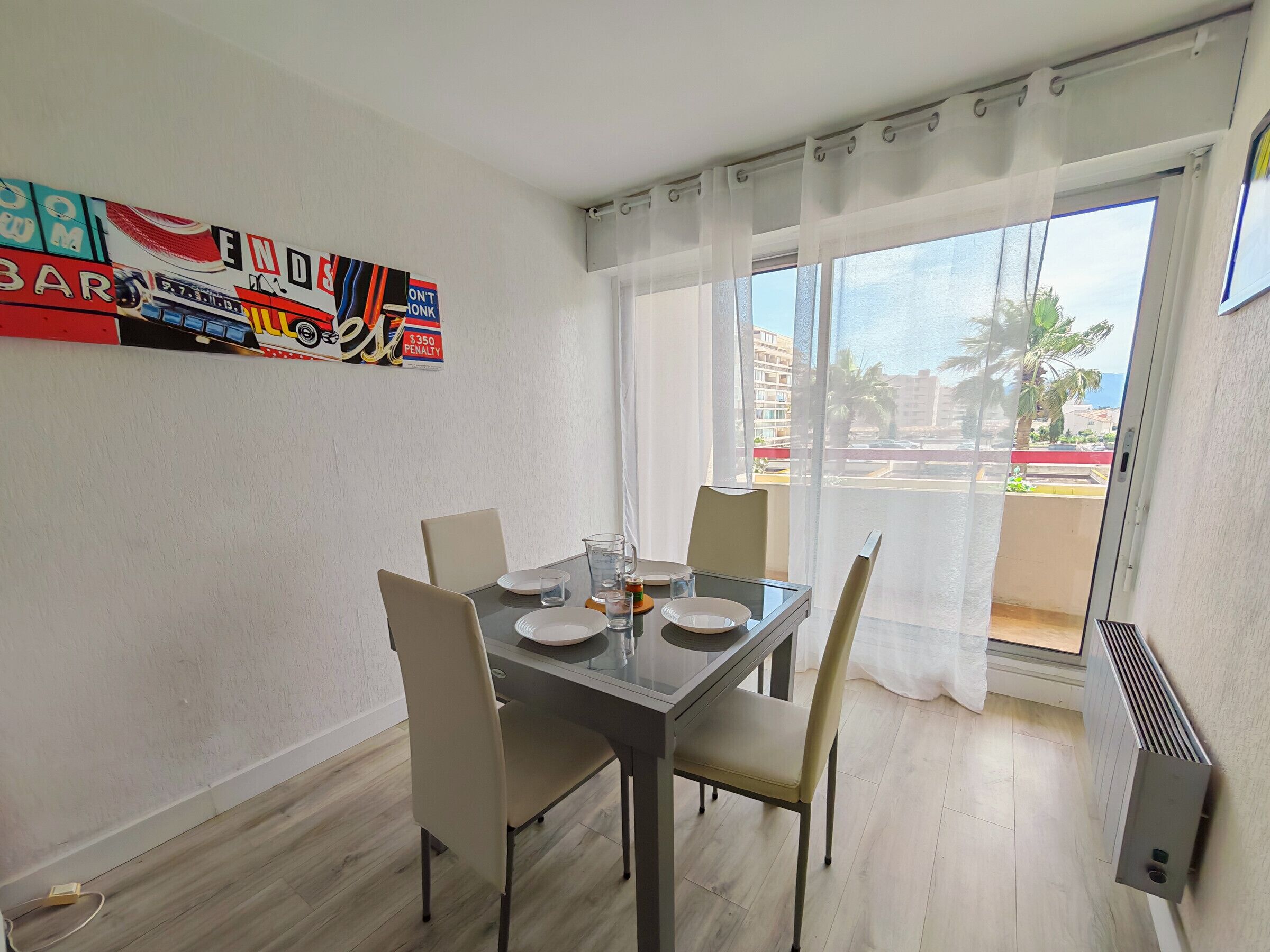 Un comedor con una mesa lista para cuatro personas, sillas, un separador de vidrio y vistas al exterior a través de una ventana con cortinas de gasa.