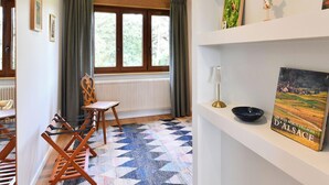 Une pièce avec une grande fenêtre, un radiateur, une chaise, une table et une étagère contenant des livres et des objets décoratifs.