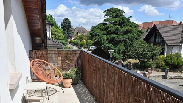 Un balcon avec un fauteuil en osier, des plantes en pot et une vue sur les maisons et les arbres des environs.