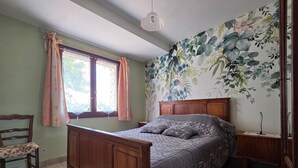 Een slaapkamer met een houten bed, een stoel, een nachtkastje, een raam met gordijnen en bloemenbehang.