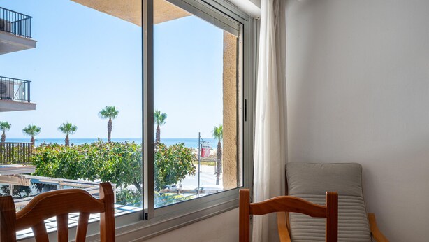 A room with a large window, wooden furniture, and a view of the beach and palm trees. - Seafront apartment, Wi-Fi included (Torredembarra)