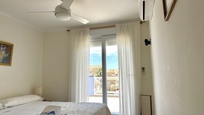 Ein Hotelzimmer mit Bett, Deckenventilator, Klimaanlage, einem Fenster mit Vorhängen und Blick auf umliegende Gebäude.