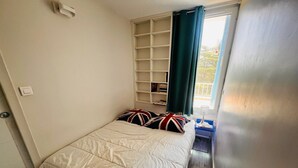 A small, neatly arranged bedroom with a bed, a shelf unit, and a window with curtains. - Apartment near the beach - 4 guests (Argelès-sur-Mer)