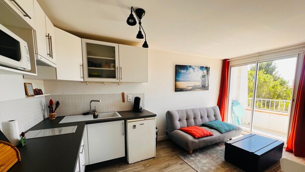 A modern kitchen with white cabinets, a black countertop, and a dining area with a grey sofa and a small table. - Apartment near the beach - 4 guests (Argelès-sur-Mer)
