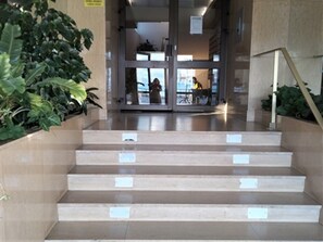 Beige Stufen führen zu einem Glastür-Eingang, im Hintergrund ist eine Person zu sehen.