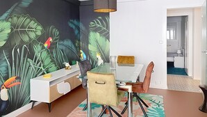 Un comedor con una mesa de vidrio, sillas, un armario blanco y un mural con aves tropicales.