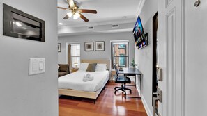 A hotel room with a bed, a desk, a ceiling fan, a TV, and a window with a city view. - Apartmint at 1149E N 3rd St Philadelphia (Philadelphia)