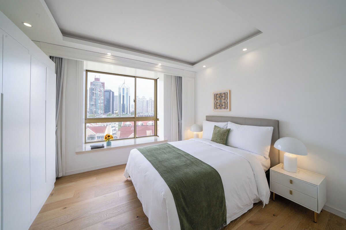 A modern bedroom with a large window, a bed with a green throw, a bedside lamp, and a nightstand.