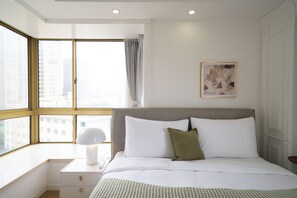 Habitación de hotel con un ventanal grande, una cama con ropa de cama blanca, una mesita de noche con lámpara y una obra de arte enmarcada en la pared.