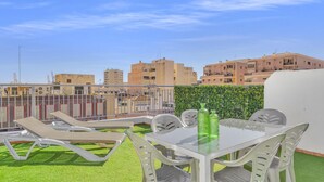 Terraço no telhado com mesa e cadeiras de vidro, um pequeno jardim com grama sintética e vista para edifícios ao redor.