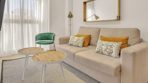 Uma sala de estar moderna com um sofá bege, duas mesas redondas, uma poltrona verde, um abajur e um espelho grande na parede.