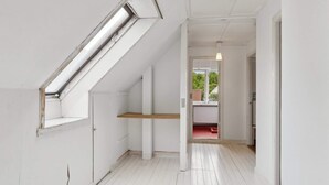 Un intérieur aux murs blancs, doté d’un plafond en pente, d’une lucarne, d’une étagère en bois et d’un couloir menant à une porte qui donne sur un paysage verdoyant.