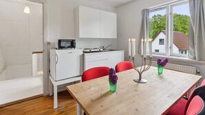 Une cuisine compacte équipée d’un four à micro-ondes, d’une cuisinière et de meubles blancs. Une table à manger avec des chaises rouges et des bougies est placée près d’une fenêtre donnant sur une maison.