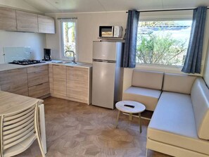 A compact kitchenette with a stove, sink, and microwave, a small round table, and a sofa by a window. - Mobile home with AC - 4 guests (Rocamadour)
