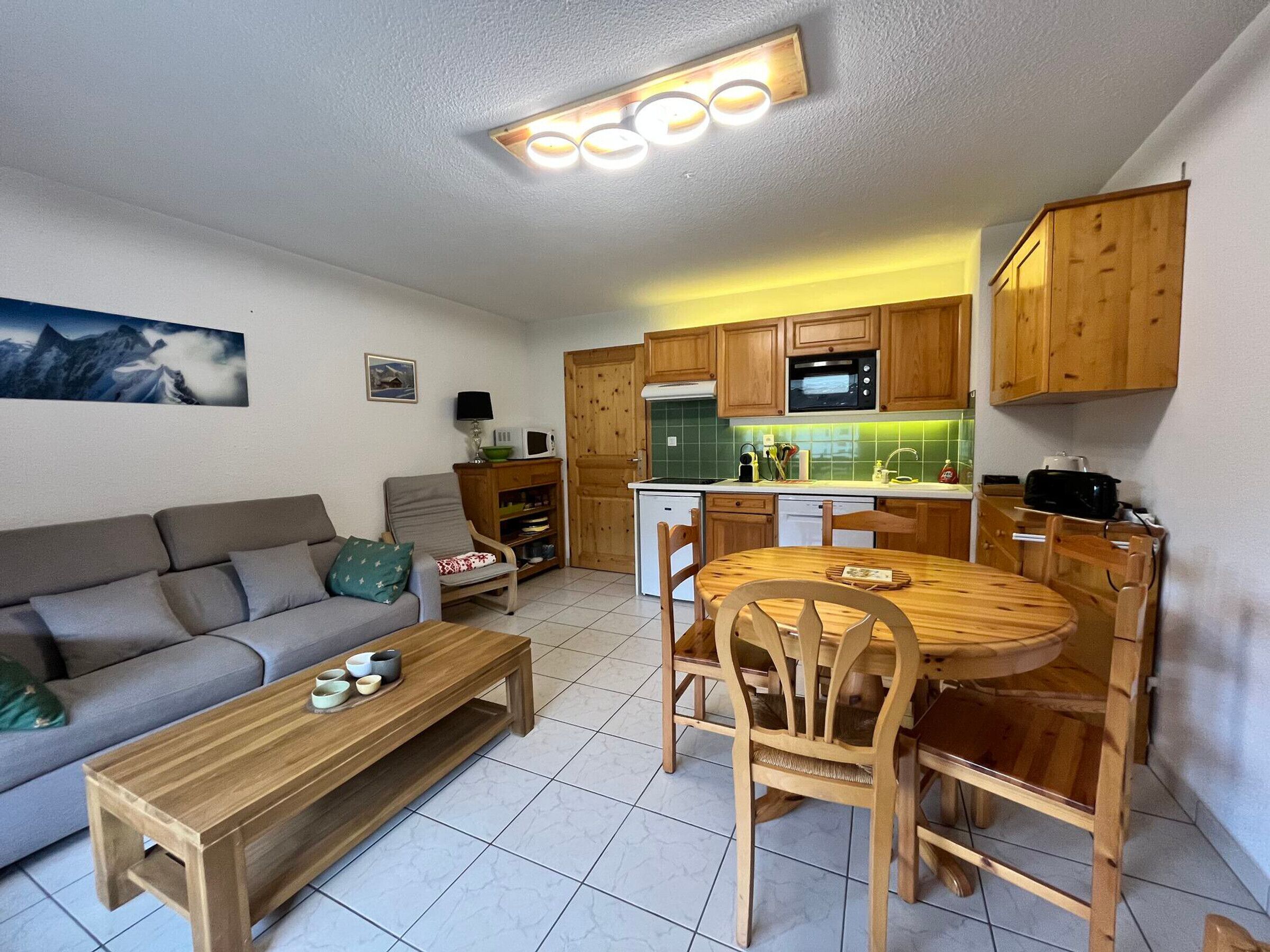 Een gezellig, compact leefruimte met een bank, een houten eettafel, stoelen, een keuken met houten kastjes, een magnetron en een kleine koelkast.