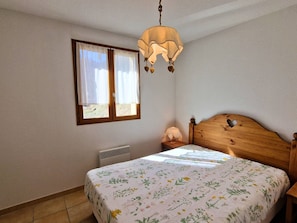 Ein Schlafzimmer mit Holzkopfende, einem Bett mit blumigem Überwurf, einem Nachttisch mit Lampe, einem Fenster mit Vorhängen und einer Deckenleuchte.